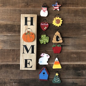 May include: Wooden HOME sign with a pumpkin and various seasonal hanging ornaments. Ornaments include a snowman, star, sunflower, shamrock, bee heart, apple, bunny, birdhouse, candy corn, and Christmas tree.