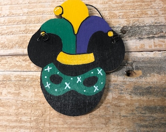 May include: A wooden Mardi Gras-themed ornament featuring a jester's hat in yellow, green, and purple, with a black base and a green mask with white crosses. The ornament is set against a rustic wooden background.