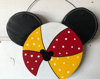 May include: Wooden Mickey Mouse-themed wall hanging. The design features black ears, a segmented bow in red, yellow, and white with white polka dots, and a black Mickey Mouse head shape in the center. A black wire loop is attached for hanging.