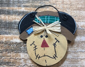 May include: A wooden scarecrow ornament with a painted face, a straw bow, and a black hat with a green plaid patch. The scarecrow has a red triangle nose and stitched mouth. A black wire loop is attached for hanging.