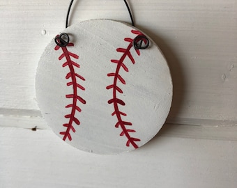 May include: A round, white wooden baseball ornament with red stitching detail. The ornament has a black wire hanger at the top. The baseball design is painted on a white background.