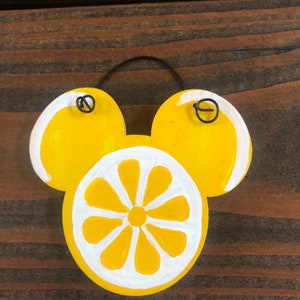 May include: A wooden lemon-themed decoration in the shape of a mouse head. The design features a yellow lemon slice with white accents and two yellow lemon halves. A black wire loop is attached for hanging.