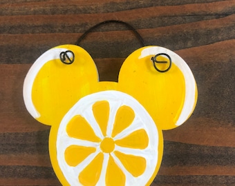 May include: A wooden lemon-themed decoration in the shape of a mouse head. The design features a yellow lemon slice with white accents and two yellow lemon halves. A black wire loop is attached for hanging.