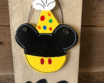 May include: Wooden sign with a black Mickey Mouse head design, featuring a yellow face with red dots, black ears, and a party hat with colorful polka dots. The sign is attached to a weathered wooden plank.
