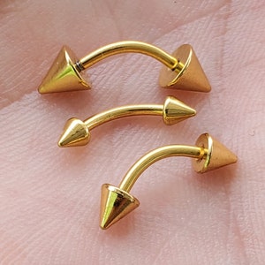 May include: Three gold-colored body piercing jewelry pieces with pointed ends. Each piece is a curved barbell with a ball on one end and a pointed end on the other.
