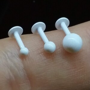 May include: Four white barbell body piercing jewelry pieces with different sized balls on the ends. The jewelry is displayed on a finger.