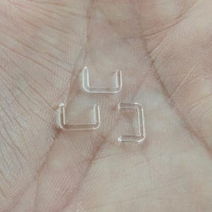Puede incluir: Tres anillos de nariz de pl&aacute;stico transparente, cada uno con forma de U, se muestran en una mano.