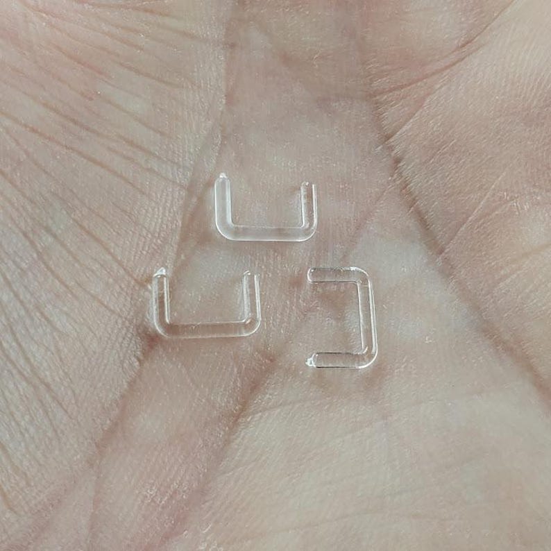 Puede incluir: Tres anillos de nariz de pl&aacute;stico transparente, cada uno con forma de U, se muestran en una mano.