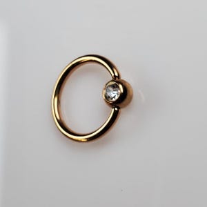 May include: Gold-toned body jewelry with a clear gem. The ring is a captive bead ring.