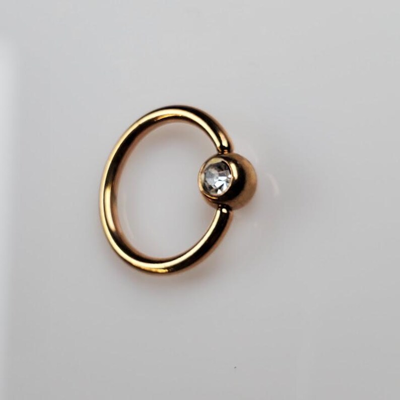 May include: Gold-toned body jewelry with a clear gem. The ring is a captive bead ring.