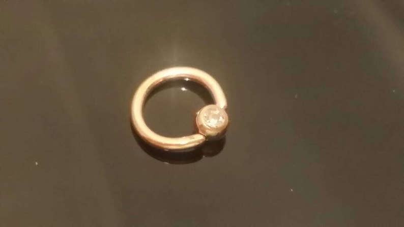 May include: A gold-toned ring with a clear gemstone set in the center. The ring is a captive bead ring, a type of body jewelry.