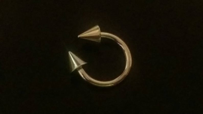May include: A gold-toned circular body piercing with two pointed ends.