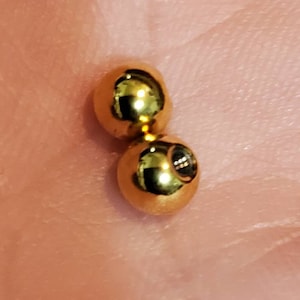 May include: Two gold-colored metal balls, one slightly larger than the other, with a small hole in the center of the smaller ball.