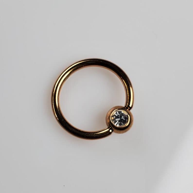 May include: A gold-toned circular body piercing with a clear gem set in a small ball at the end.