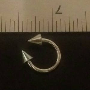 May include: Silver body piercing with two pointed ends. The piercing is about 1 inch in diameter.