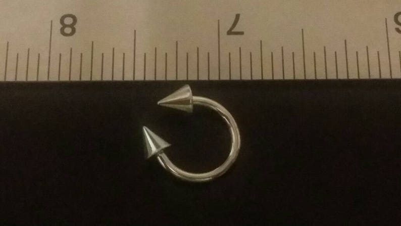 May include: Silver body piercing with two pointed ends. The piercing is about 1 inch in diameter.