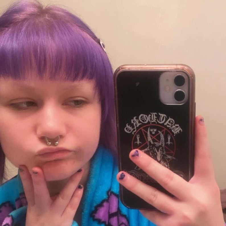 May include: A person with purple hair is holding a black phone case with a pentagram and the words "Evil Dead" printed on it. The phone case has a gold trim.