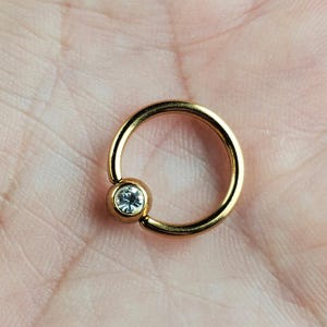 May include: Gold body piercing ring with a clear gem in the center.