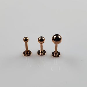 May include: Three gold-colored barbell studs with different sized balls. The balls are round and smooth. The studs are arranged in order of size from smallest to largest.