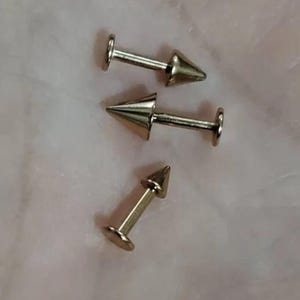 May include: Three gold-colored body piercing studs with a pointed tip. The studs are designed for cartilage piercings.