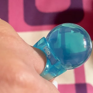 以下が含まれることがあります： 大きな球状のトップが付いた、透明な青色のリング。リングのバンドには「MIR」の文字がプリントされています。リングはピンクと紫の背景に表示されています。