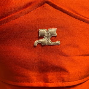 Pu&ograve; includere: Primo piano di un capo d'abbigliamento arancione con un logo bianco. Il tessuto ha un aspetto strutturato. Il logo &egrave; un design stilizzato, forse un emblema del marchio, ricamato sul capo.