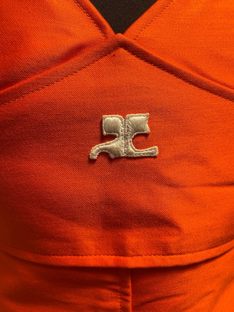Pu&ograve; includere: Primo piano di un capo d'abbigliamento arancione con un logo bianco. Il tessuto ha un aspetto strutturato. Il logo &egrave; un design stilizzato, forse un emblema del marchio, ricamato sul capo.