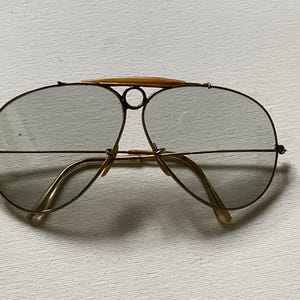 Może przedstawiać: Okulary przeciwsłoneczne typu aviator z cienkimi, złotymi metalowymi oprawkami i lekko przyciemnianymi szkłami. Konstrukcja obejmuje wyraźny g&oacute;rny mostek z centralnym okrągłym detalem oraz jasny element ozdobny umieszczony na g&oacute;rze oprawek.