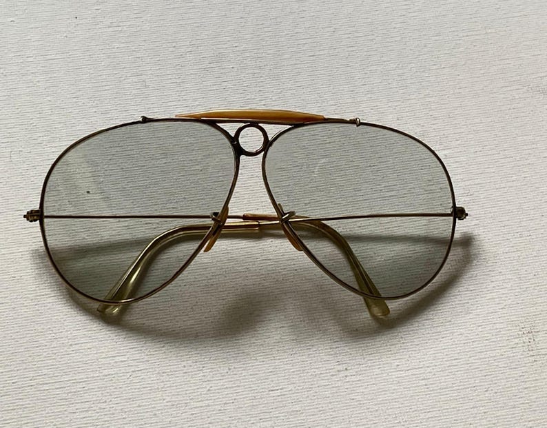 Może przedstawiać: Okulary przeciwsłoneczne typu aviator z cienkimi, złotymi metalowymi oprawkami i lekko przyciemnianymi szkłami. Konstrukcja obejmuje wyraźny g&oacute;rny mostek z centralnym okrągłym detalem oraz jasny element ozdobny umieszczony na g&oacute;rze oprawek.