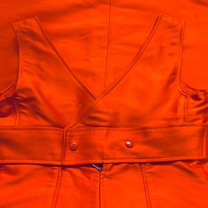 Pu&ograve; includere: Gilet arancione brillante con scollo a V e cintura avvolgente. Il gilet ha una chiusura a bottoni doppiopetto e una cerniera. Il tessuto ha una leggera lucentezza e il capo &egrave; posto su uno sfondo arancione abbinato.