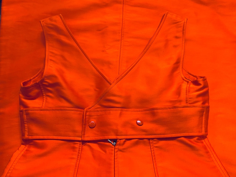 Pu&ograve; includere: Gilet arancione brillante con scollo a V e cintura avvolgente. Il gilet ha una chiusura a bottoni doppiopetto e una cerniera. Il tessuto ha una leggera lucentezza e il capo &egrave; posto su uno sfondo arancione abbinato.
