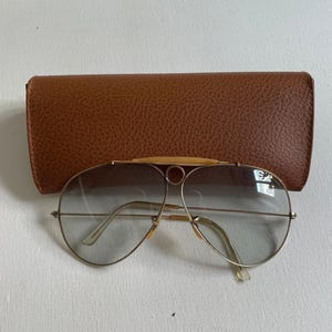 Może przedstawiać: Okulary przeciwsłoneczne typu aviator z cienkimi złotymi metalowymi oprawkami, szarymi soczewkami z gradientem oraz charakterystycznym detalem na g&oacute;rnej belce z małym okrągłym elementem mostka. Okulary są prezentowane w teksturowanym brązowym etui.