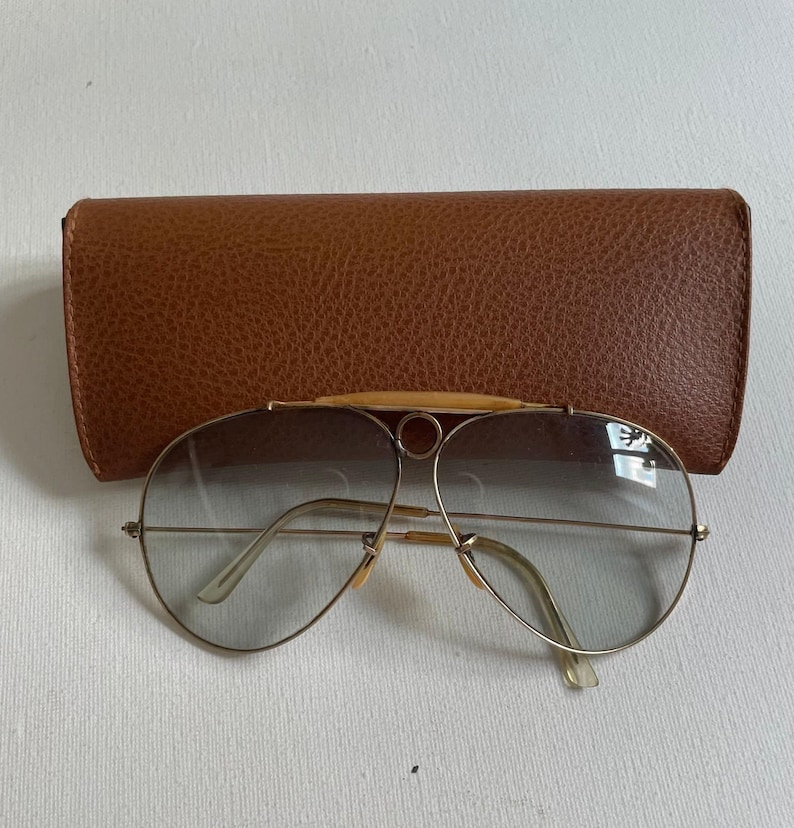 Może przedstawiać: Okulary przeciwsłoneczne typu aviator z cienkimi złotymi metalowymi oprawkami, szarymi soczewkami z gradientem oraz charakterystycznym detalem na g&oacute;rnej belce z małym okrągłym elementem mostka. Okulary są prezentowane w teksturowanym brązowym etui.