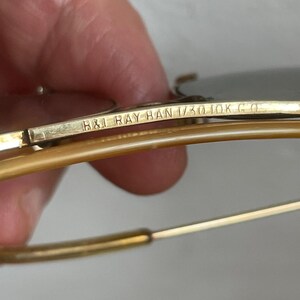 Może przedstawiać: Okulary przeciwsłoneczne w kolorze złotym z metalową poprzeczką, na kt&oacute;rej wygrawerowano napis B&L RAY BAN 1/30 10K G O. Oprawka posiada cienkie, złote zauszniki z drutu oraz jasnobrązowe, p&oacute;łprzezroczyste końc&oacute;wki z tworzywa w stylu lotniczym.