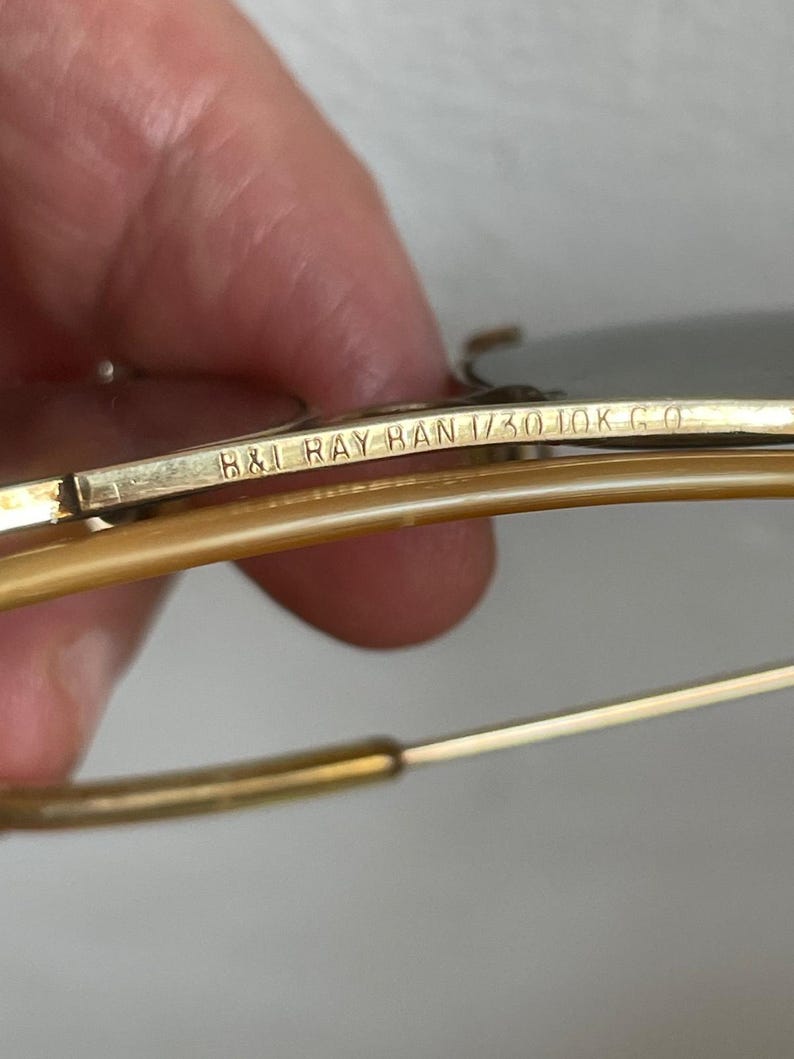 Może przedstawiać: Okulary przeciwsłoneczne w kolorze złotym z metalową poprzeczką, na kt&oacute;rej wygrawerowano napis B&L RAY BAN 1/30 10K G O. Oprawka posiada cienkie, złote zauszniki z drutu oraz jasnobrązowe, p&oacute;łprzezroczyste końc&oacute;wki z tworzywa w stylu lotniczym.