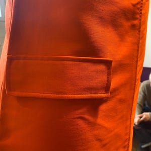 Pu&ograve; includere: Primo piano di un tessuto arancione vibrante con un dettaglio di tasca rettangolare. La tasca &egrave; delineata da una cucitura a contrasto, che aggiunge un sottile elemento di texture al materiale liscio e tinta unita. L'immagine evidenzia la costruzione sartoriale.