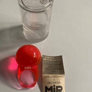 以下が含まれることがあります： 赤いプラスチック製のリングで、球状のトップと「MiR」の文字が透明なプラスチック容器の隣に表示されています。イタリア語のテキストと「MiR」のロゴが入った小さなカードも付いています。リングはユニークなアクセサリーです。