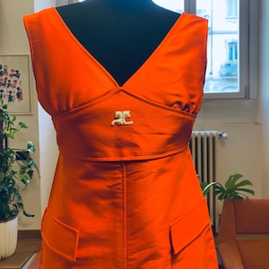 Pu&ograve; includere: Abito arancione brillante con scollo a V e un emblema dorato. L'abito presenta un corpetto aderente, una vita definita e due tasche anteriori. &Egrave; esposto su un manichino nero.