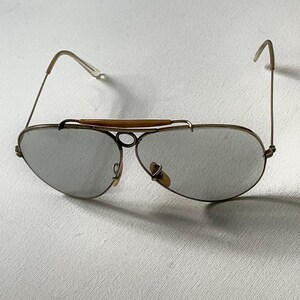 Może przedstawiać: Okulary przeciwsłoneczne typu aviator z cienkimi, złotymi metalowymi oprawkami, szarymi soczewkami w kształcie łezki oraz charakterystyczną beżową poprzeczką nad mostkiem. Zauszniki są smukłe, z przezroczystymi końc&oacute;wkami i jasnobeżowymi noskami.