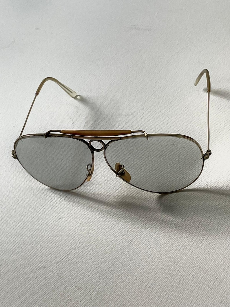 Może przedstawiać: Okulary przeciwsłoneczne typu aviator z cienkimi, złotymi metalowymi oprawkami, szarymi soczewkami w kształcie łezki oraz charakterystyczną beżową poprzeczką nad mostkiem. Zauszniki są smukłe, z przezroczystymi końc&oacute;wkami i jasnobeżowymi noskami.