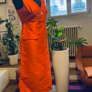 Pu&ograve; includere: Abito arancione brillante senza maniche con scollo a V e una piccola tasca. L'abito &egrave; esposto su un manichino nero. Un piccolo logo &egrave; visibile sul petto. L'abito &egrave; lungo circa 140 cm.