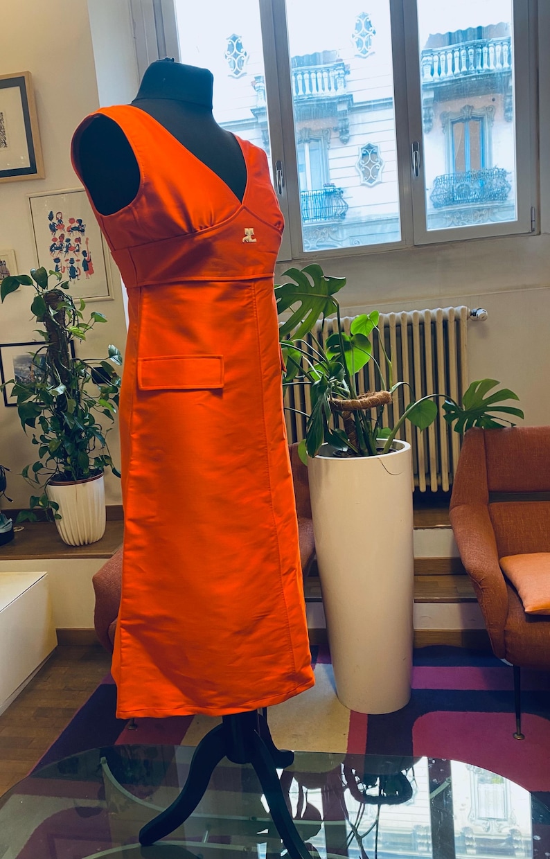 Pu&ograve; includere: Abito arancione brillante senza maniche con scollo a V e una piccola tasca. L'abito &egrave; esposto su un manichino nero. Un piccolo logo &egrave; visibile sul petto. L'abito &egrave; lungo circa 140 cm.