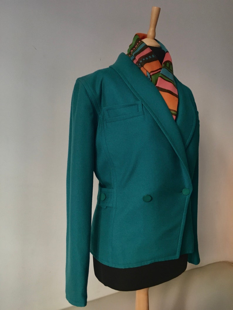 May include: A teal double-breasted blazer with a long sleeve and two buttons. The blazer has a pocket on the left side and is worn on a mannequin with a colorful patterned scarf.