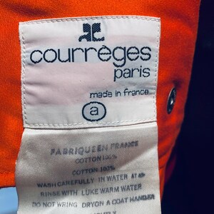 Pu&ograve; includere: Primo piano di un'etichetta bianca con la scritta "courr&egrave;ges paris made in france" in nero. L'etichetta &egrave; cucita su un tessuto arancione. Sono visibili anche le istruzioni di lavaggio.