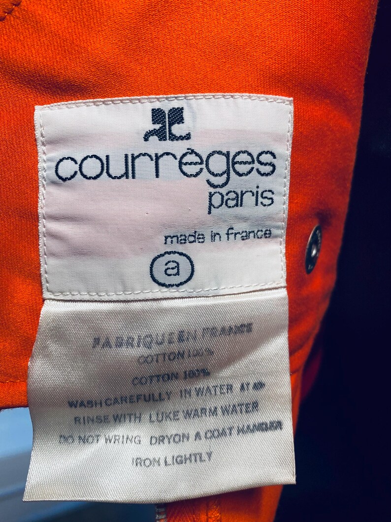 Pu&ograve; includere: Primo piano di un'etichetta bianca con la scritta "courr&egrave;ges paris made in france" in nero. L'etichetta &egrave; cucita su un tessuto arancione. Sono visibili anche le istruzioni di lavaggio.