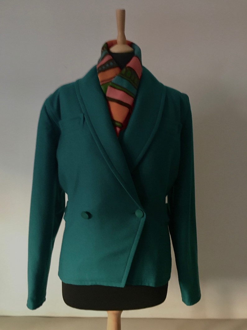 May include: A teal green blazer with a double breasted closure and long sleeves. The blazer has a shawl collar and is worn on a mannequin with a patterned scarf.