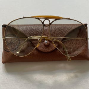 Może przedstawiać: Złote okulary przeciwsłoneczne typu aviator z metalu z wyraźnym g&oacute;rnym paskiem z ż&oacute;łtym akcentem, umieszczone na brązowym, fakturowanym sk&oacute;rzanym etui z zapięciem na zatrzask. Soczewki są przyciemniane, a oprawka ma konstrukcję z podw&oacute;jnym mostkiem.