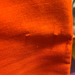 Pu&ograve; includere: Primo piano di un tessuto arancione vibrante, che mostra la trama e l'ordito del materiale. Il colore brillante domina la cornice, con sottili variazioni di tono e un bordo leggermente sfilacciato.