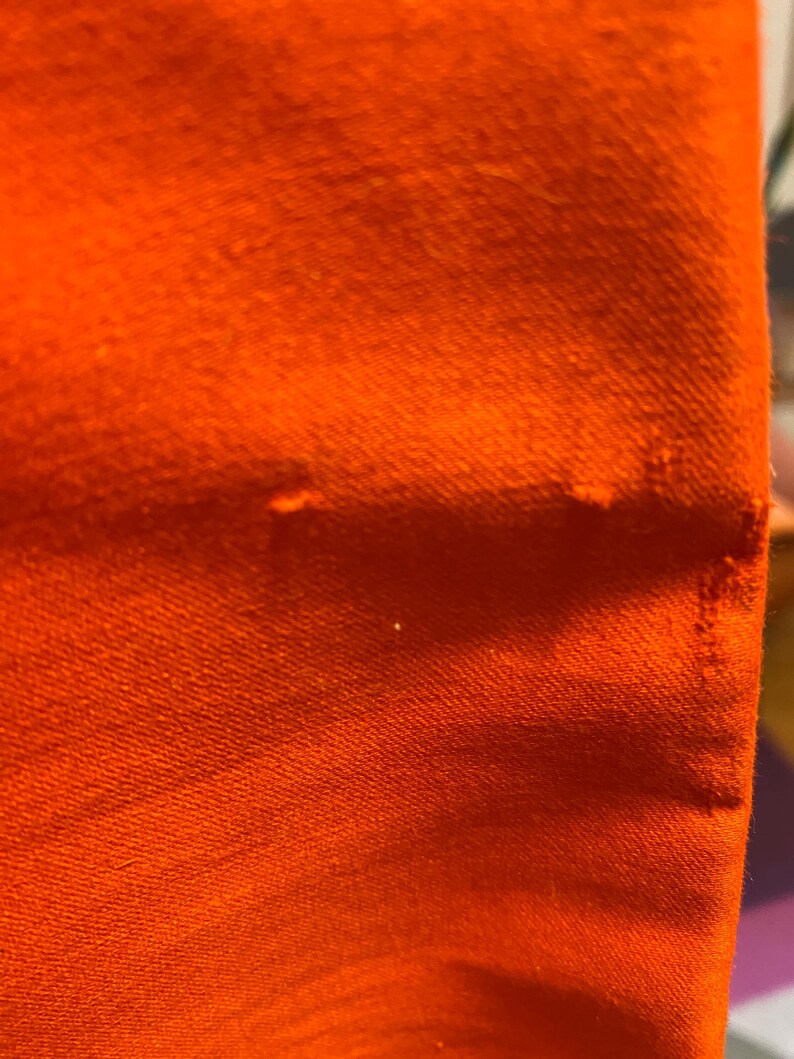 Pu&ograve; includere: Primo piano di un tessuto arancione vibrante, che mostra la trama e l'ordito del materiale. Il colore brillante domina la cornice, con sottili variazioni di tono e un bordo leggermente sfilacciato.
