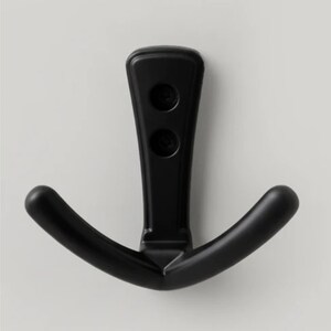 May include: A black, double-hook wall mount. The hook has a vertical stem with two circular indentations and two curved hooks extending outwards. The wall mount is made of a matte black material.